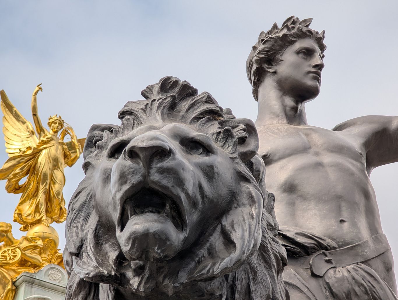 Groupe de statues devant Buckingham Palace : une tête de lion au premier plan, un guerrier derrière et une victoire ailée dorée en arrière plan.